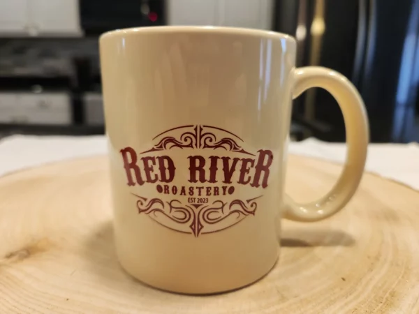 Cream colored coffee mug with maroon Red River Roastery logo.