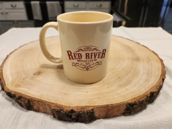 Cream colored coffee mug with maroon Red River Roastery logo, and mug handle to the left.