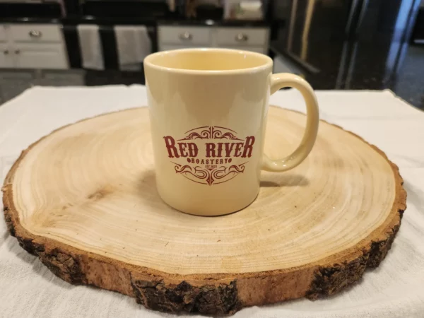 Cream colored coffee mug with maroon Red River Roastery logo, and mug handle to the right.