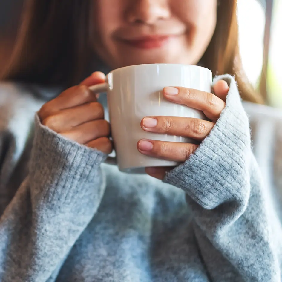 Young woman in a sweater smiling and holding a cup of fresh coffee.